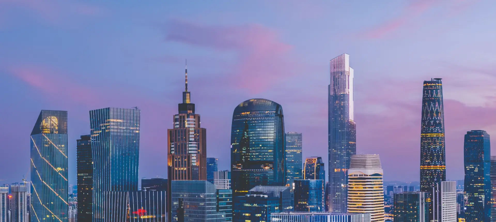 Panoramic view of a city skyline at dusk, showcasing multiple illuminated skyscrapers against a purple and blue sky.
