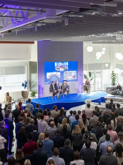 Large corporate event with speakers on stage addressing a crowded audience in a modern office space.