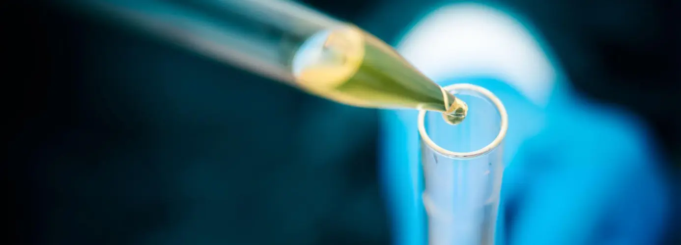 Close-up of a liquid droplet falling from a pipette into a small metal tube, with blurred blue background.