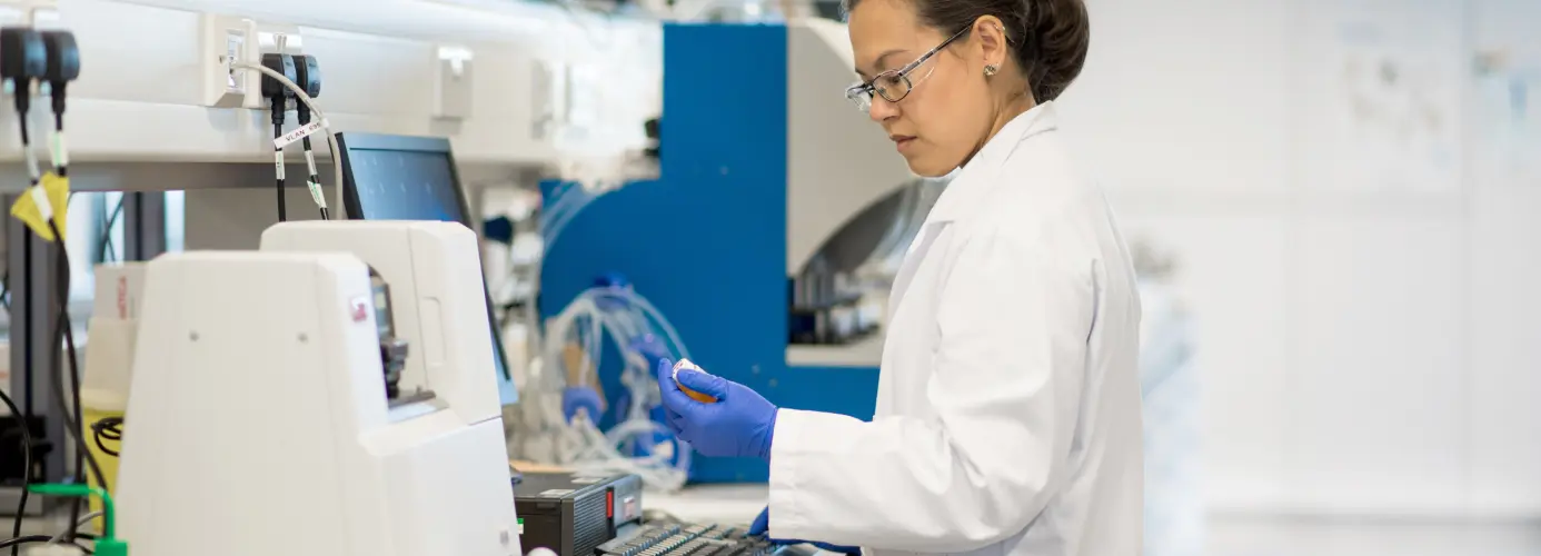A scientist in a lab coat and gloves holding a small bottle