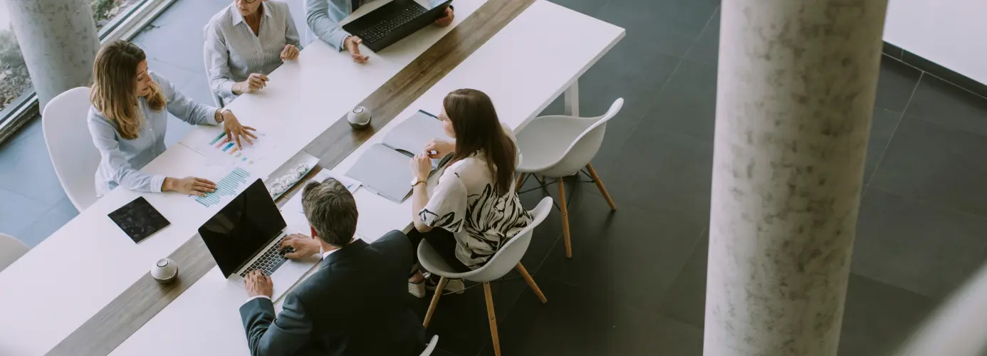Overhead view of a business meeting with five people seated around a white table, using laptops and discussing documents.