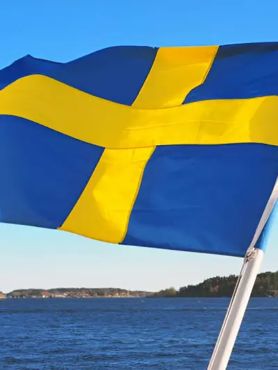 Swedish flag waving in the foreground with a lake and forested shoreline in the background.