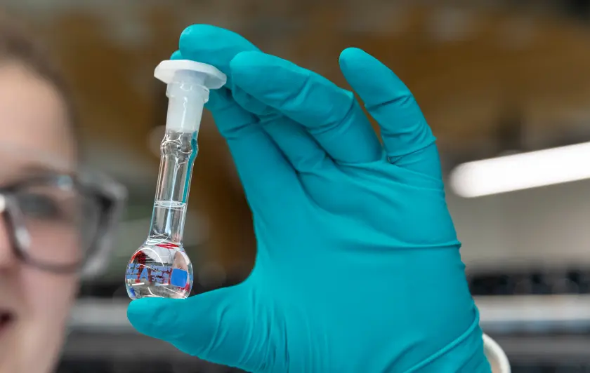 Hand in teal glove holding a small glass vial with liquid inside a laboratory setting
