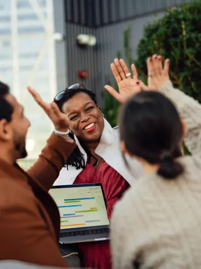 Enthusiastic team meeting with a smiling woman in the center and colleagues raising hands in agreement, laptop displaying data visible.