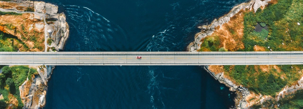 Aerial view of a bridge spanning a deep blue river gorge with rocky cliffs and green vegetation on both sides.