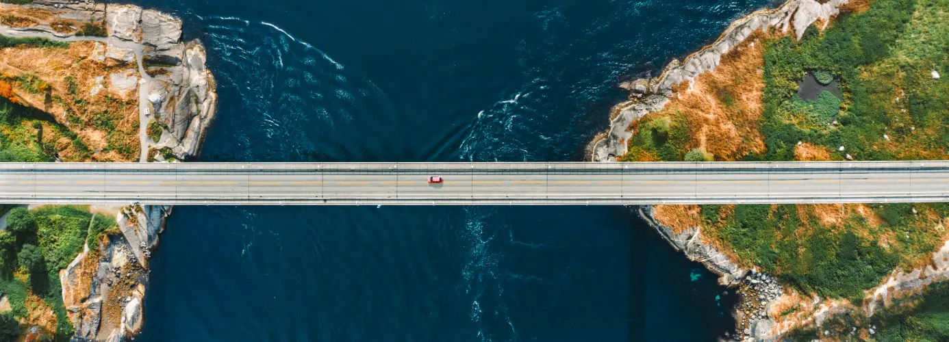 Aerial view of a bridge spanning a deep blue river gorge with rocky cliffs and green vegetation on both sides.