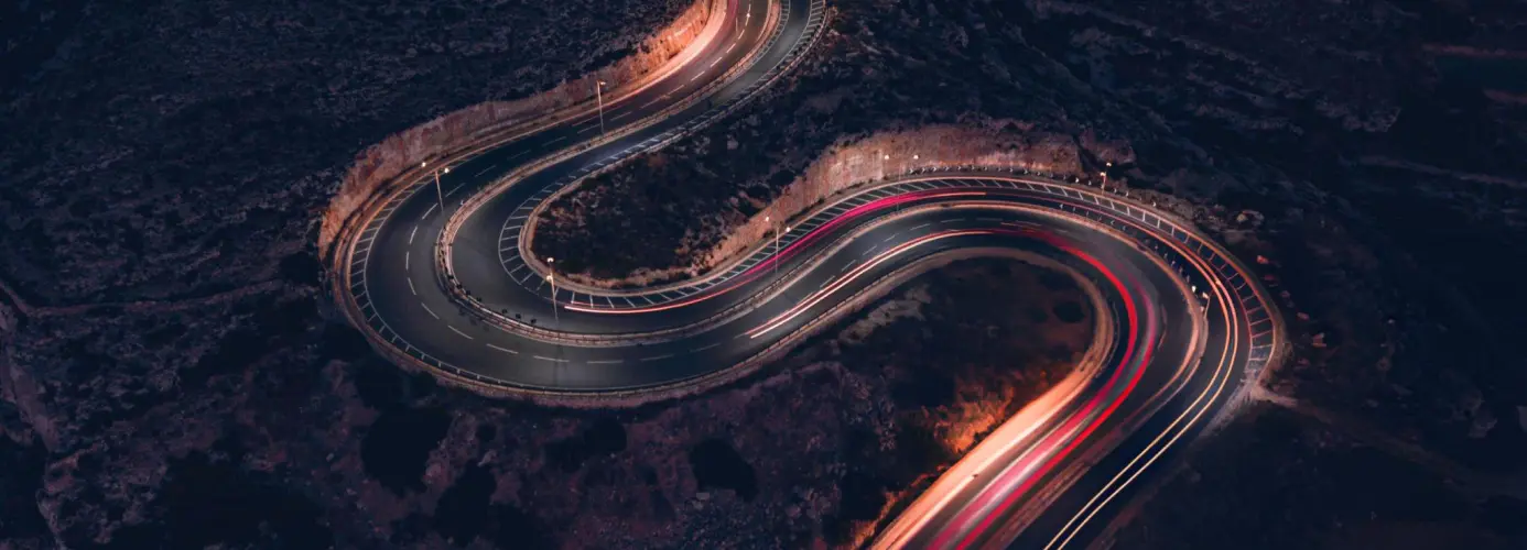 A winding mountain road at night, with long exposure light trails from vehicles creating vibrant streaks of red and white against the dark landscape.